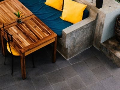 Outdoor sofa with blue covering and yellow cushions, next to a wooden reclaimed table, on grey patio paving slabs