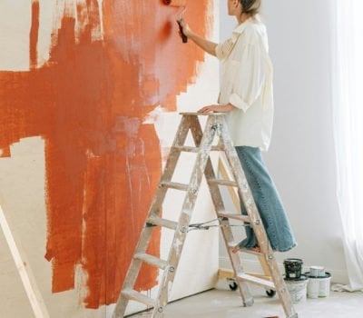 Woman on a stepladder painting a wall a burnt orange colour with a paint roller