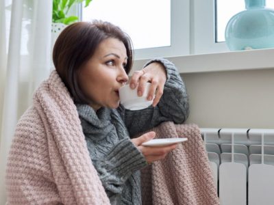 Lady staying warm by a radiator warpped in a blanket and drinking from a cup