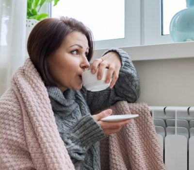Lady staying warm by a radiator warpped in a blanket and drinking from a cup