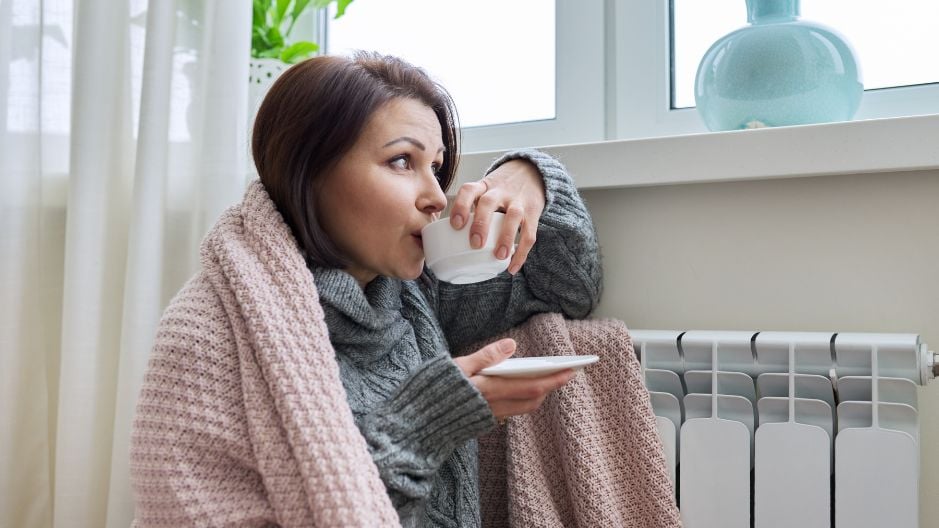 Lady staying warm by a radiator warpped in a blanket and drinking from a cup